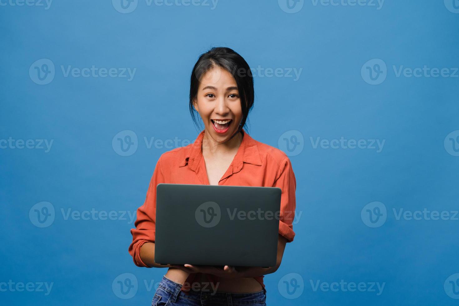 surpreendeu a jovem asiática usando laptop com expressão positiva, sorriso largo, vestido com roupas casuais e olhando para a câmera sobre fundo azul. feliz adorável feliz mulher alegra sucesso. foto