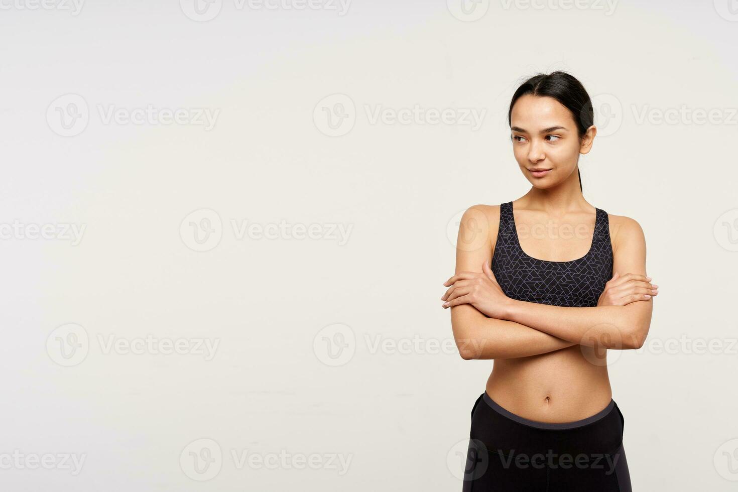 positivo jovem bonita Sombrio cabelos desportivo senhora guardando dela mãos cruzado enquanto olhando pensativamente aparte, em pé sobre branco fundo dentro Atlético vestem foto