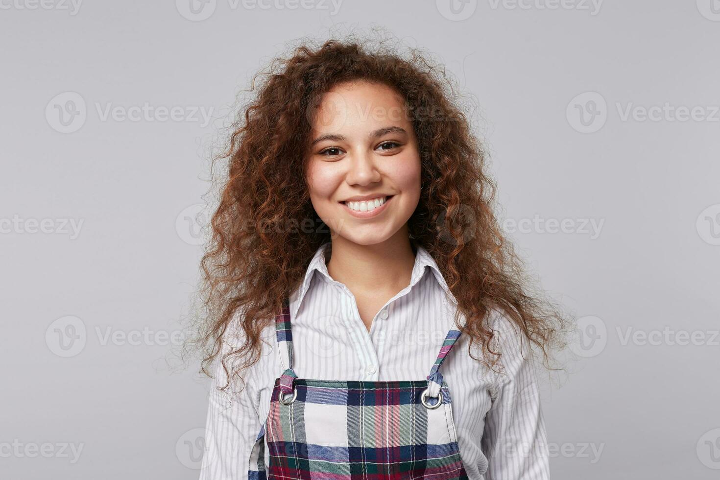 retrato do feliz jovem atraente Castanho cabelos encaracolado senhora ser dentro agradável humor e sorridente alegremente às Câmera, isolado sobre cinzento fundo dentro elegante vestem foto