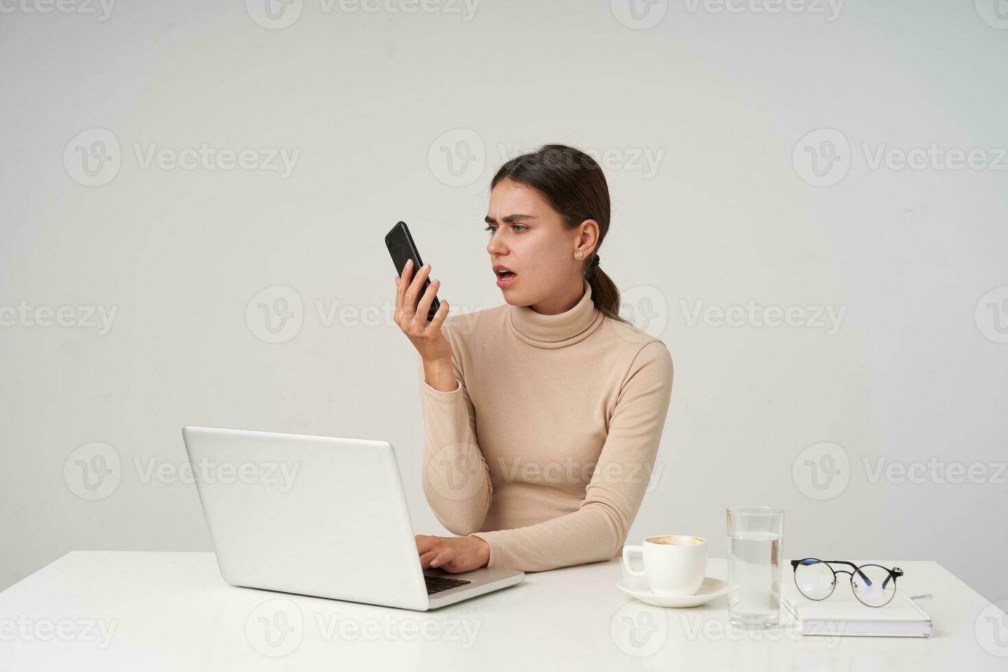 interior tiro do descontente jovem morena senhora olhando em aparelho portátil com beicinho e fazendo careta dela face, guardando mão em teclado enquanto sentado sobre branco fundo foto