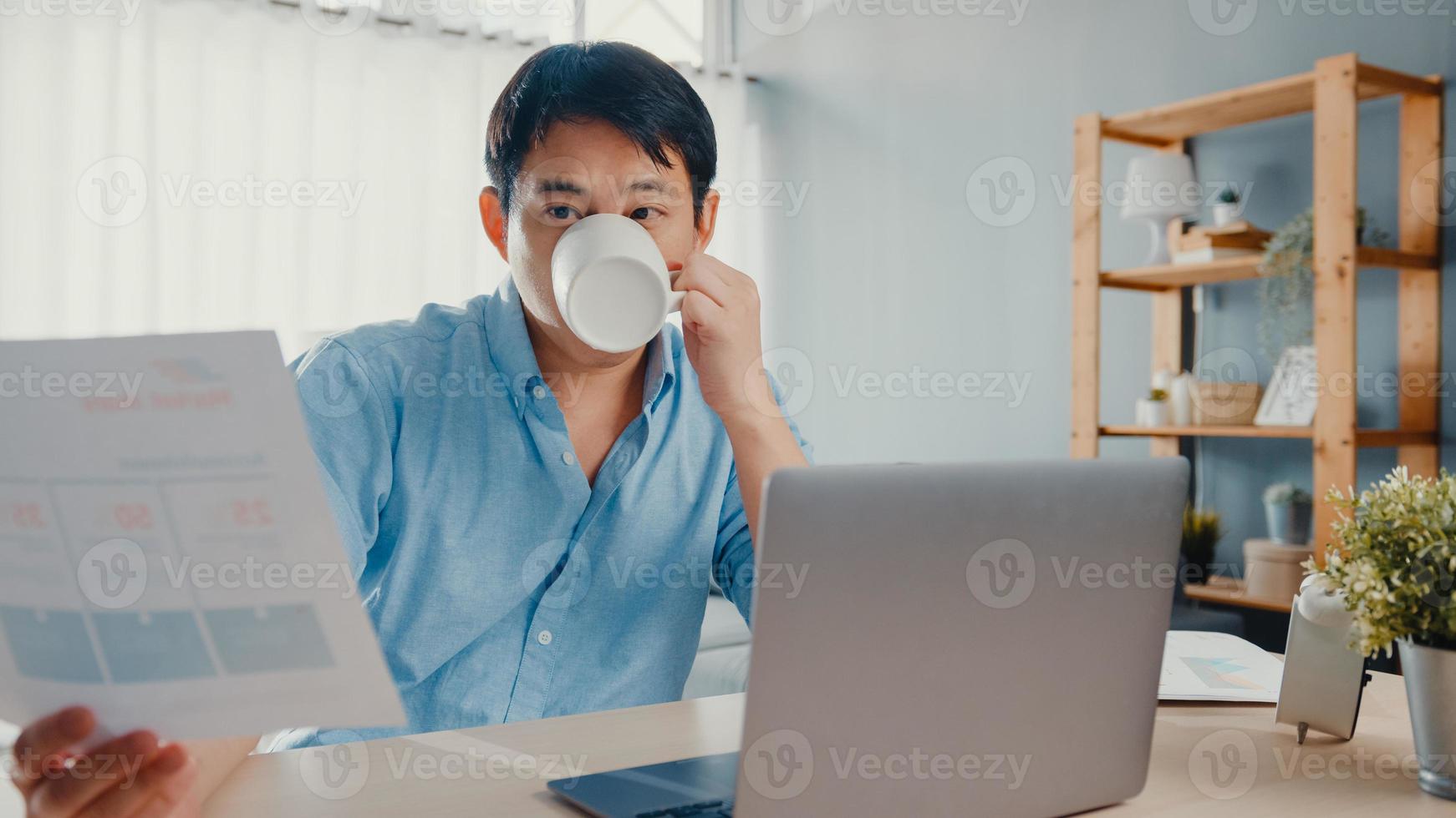 freelance asiático cara casual wear usando laptop e beber café na sala de estar em casa. trabalho de casa, trabalho remoto, educação a distância, distanciamento social, quarentena para prevenção do vírus corona. foto