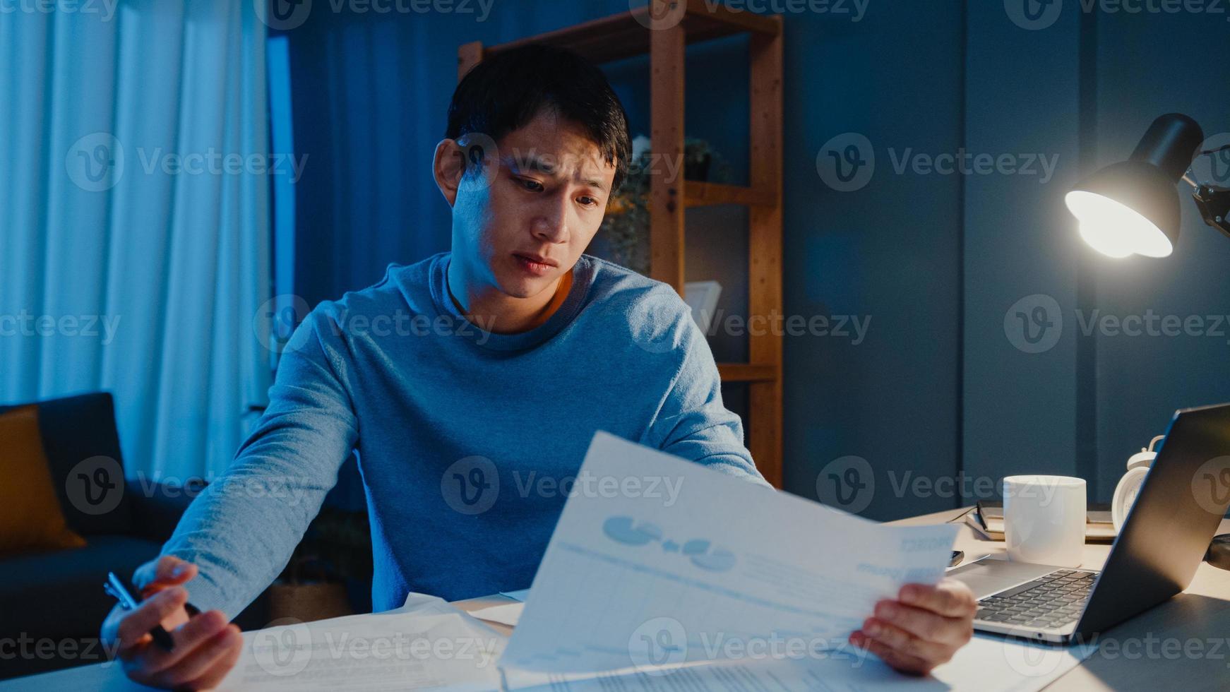empresário freelance asiático foco tipo de trabalho no computador portátil ocupado com gráfico cheio de papelada na mesa na sala de estar em casa horas extras à noite, trabalhar em casa durante o conceito de pandemia covid-19. foto