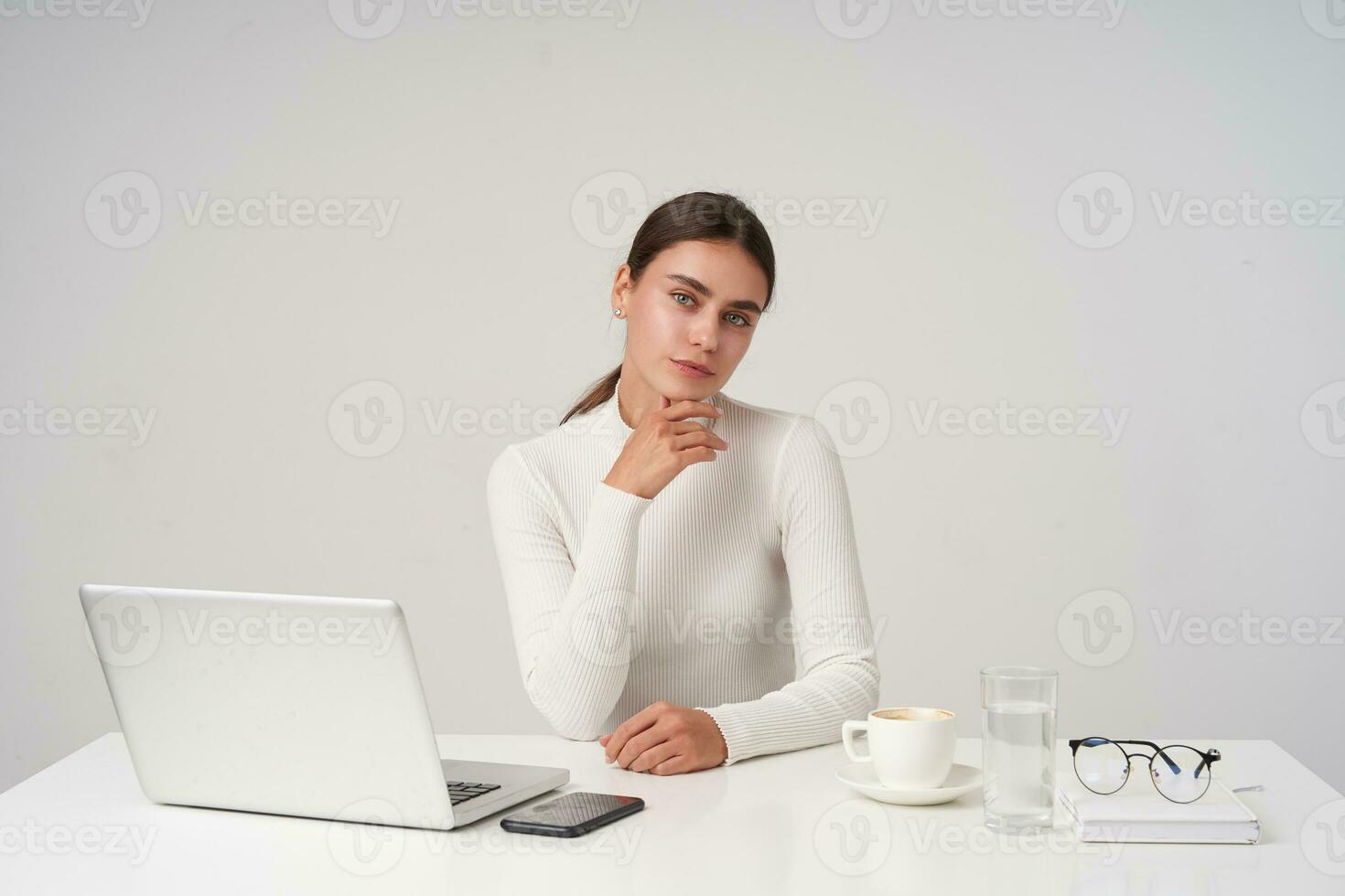 positivo jovem encantador morena senhora segurando dela queixo em elevado mão e sorridente levemente enquanto olhando às Câmera, vestindo branco tricotado Gola polo enquanto posando sobre branco fundo foto