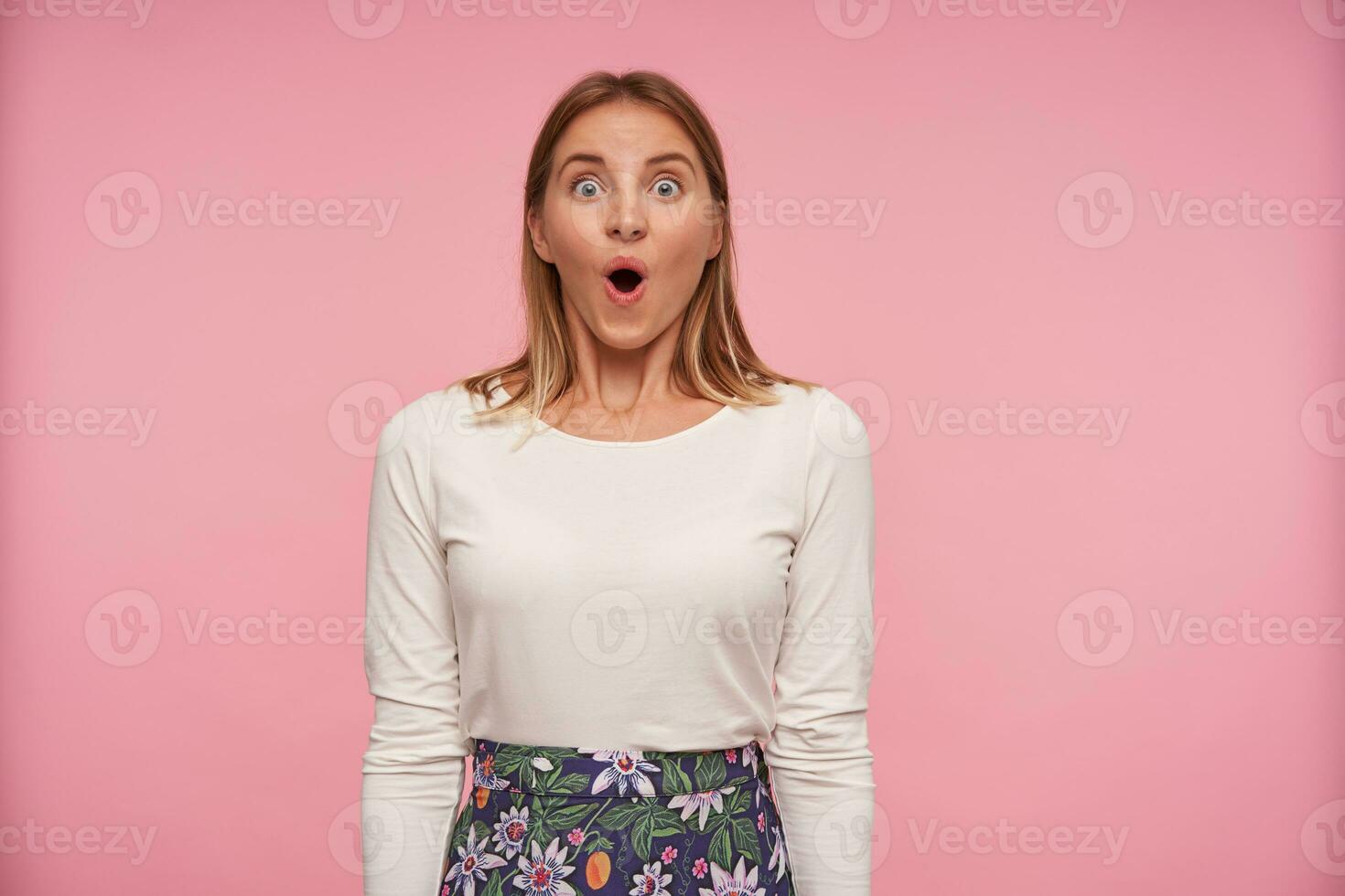 surpreso bonita jovem fêmea com Loiras cabelo vestindo branco blusa e floresceu saia enquanto em pé sobre Rosa fundo, arredondamento olhos e levantando sobrancelhas surpreendentemente enquanto olhando para Câmera foto