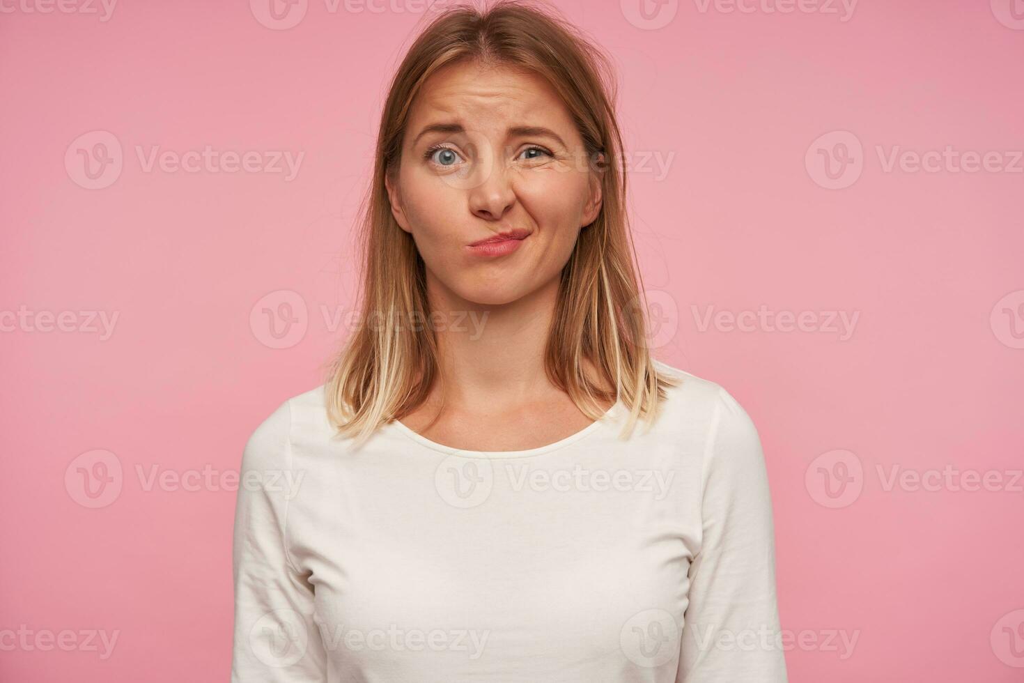 confuso de olhos azuis bonita fêmea com Loiras cabelo olhando às Câmera com confuso face e fazendo careta, vestido dentro casual vestem enquanto posando sobre Rosa fundo foto