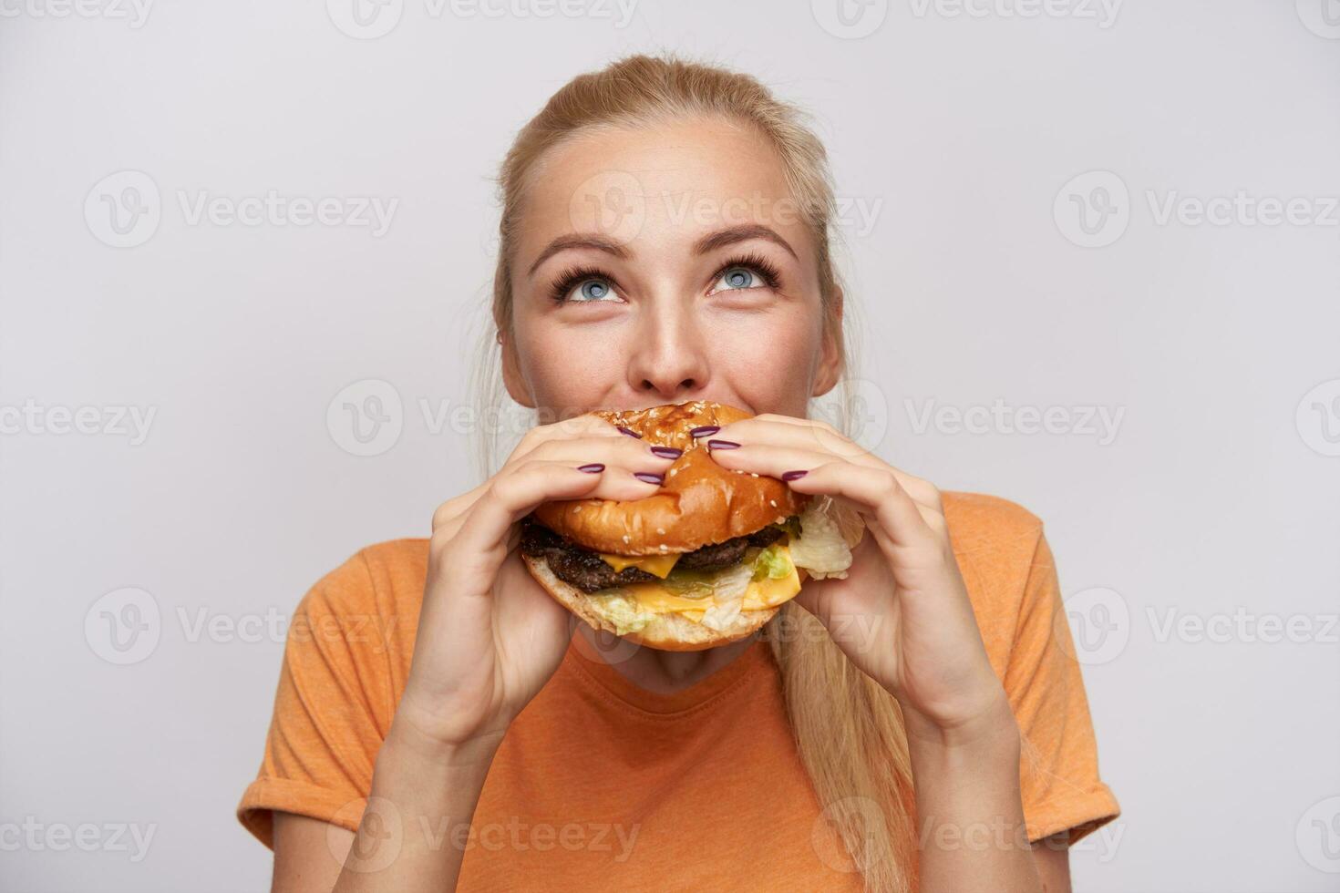 retrato do satisfeito jovem adorável Loiras mulher com casual Penteado comendo fresco Hamburger com ótimo apetite e olhando alegremente para cima, posando sobre branco fundo foto