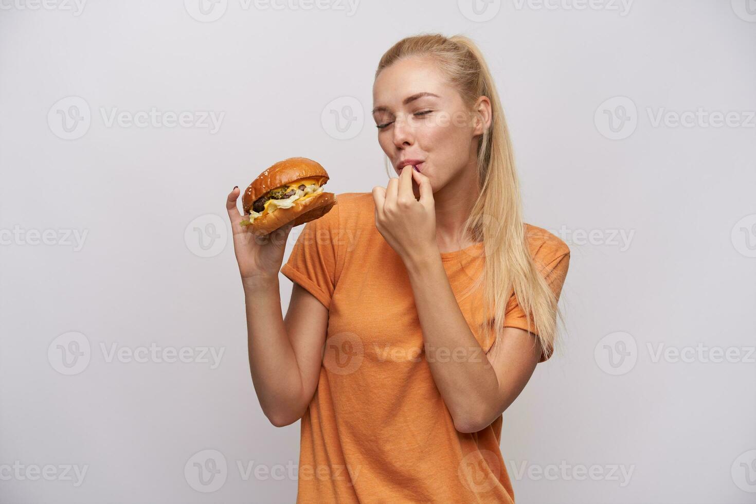 interior tiro do satisfeito jovem grandes cabelos Loiras fêmea com rabo de cavalo Penteado guardando dela olhos fechadas enquanto degustação grande fresco hamburguer dentro dela mão, isolado sobre branco fundo foto