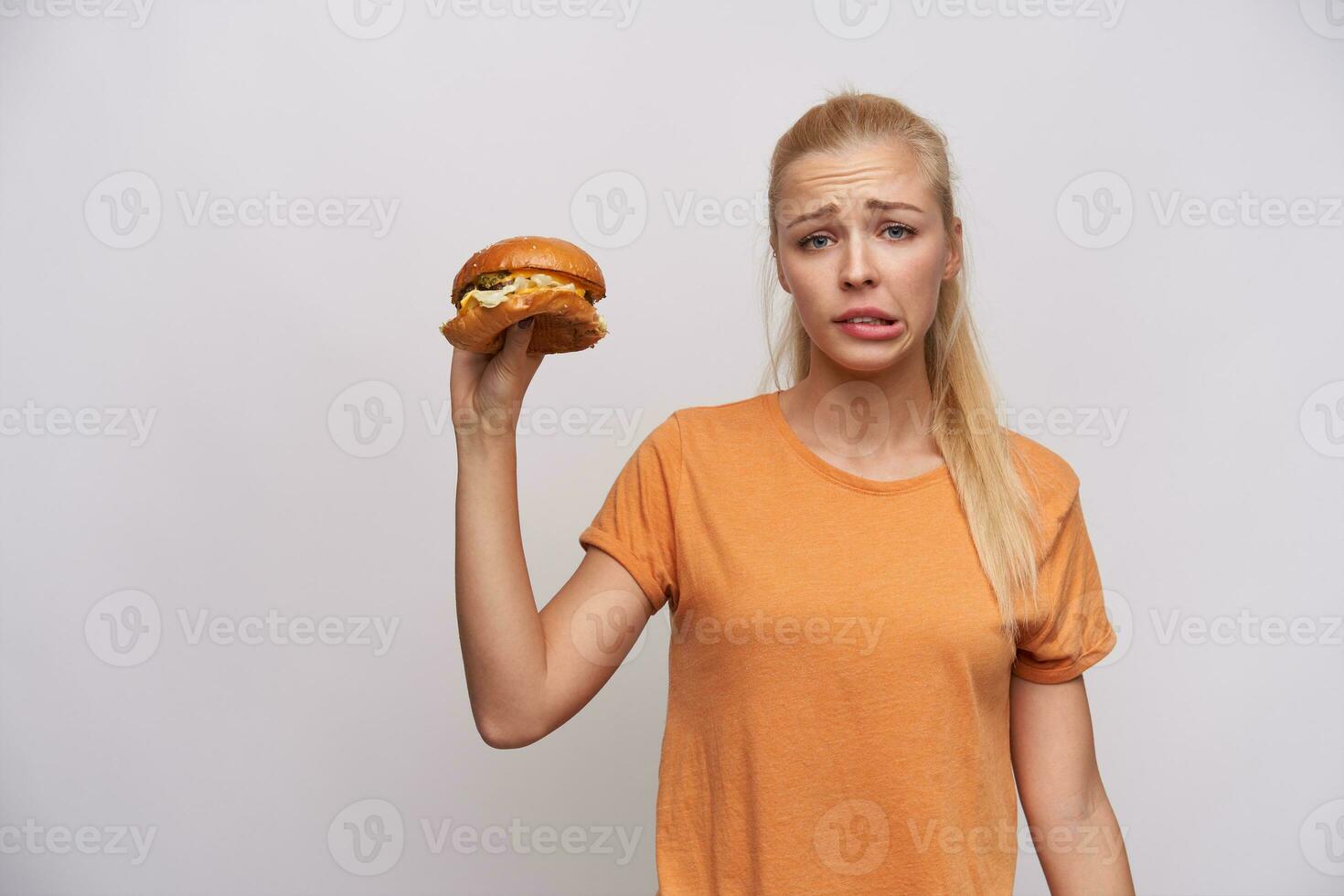 descontente jovem bonita grandes cabelos Loiras mulher com rabo de cavalo Penteado franzindo a testa dela face com beicinho enquanto segurando lixo Comida dentro elevado mão, em pé contra branco fundo foto