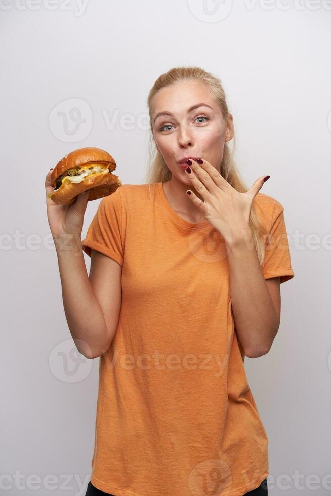 alegre jovem bonita Loiras mulher com casual Penteado olhando positivamente às Câmera e guardando mão em dela boca enquanto degustação grande fresco hambúrguer, em pé contra branco fundo foto