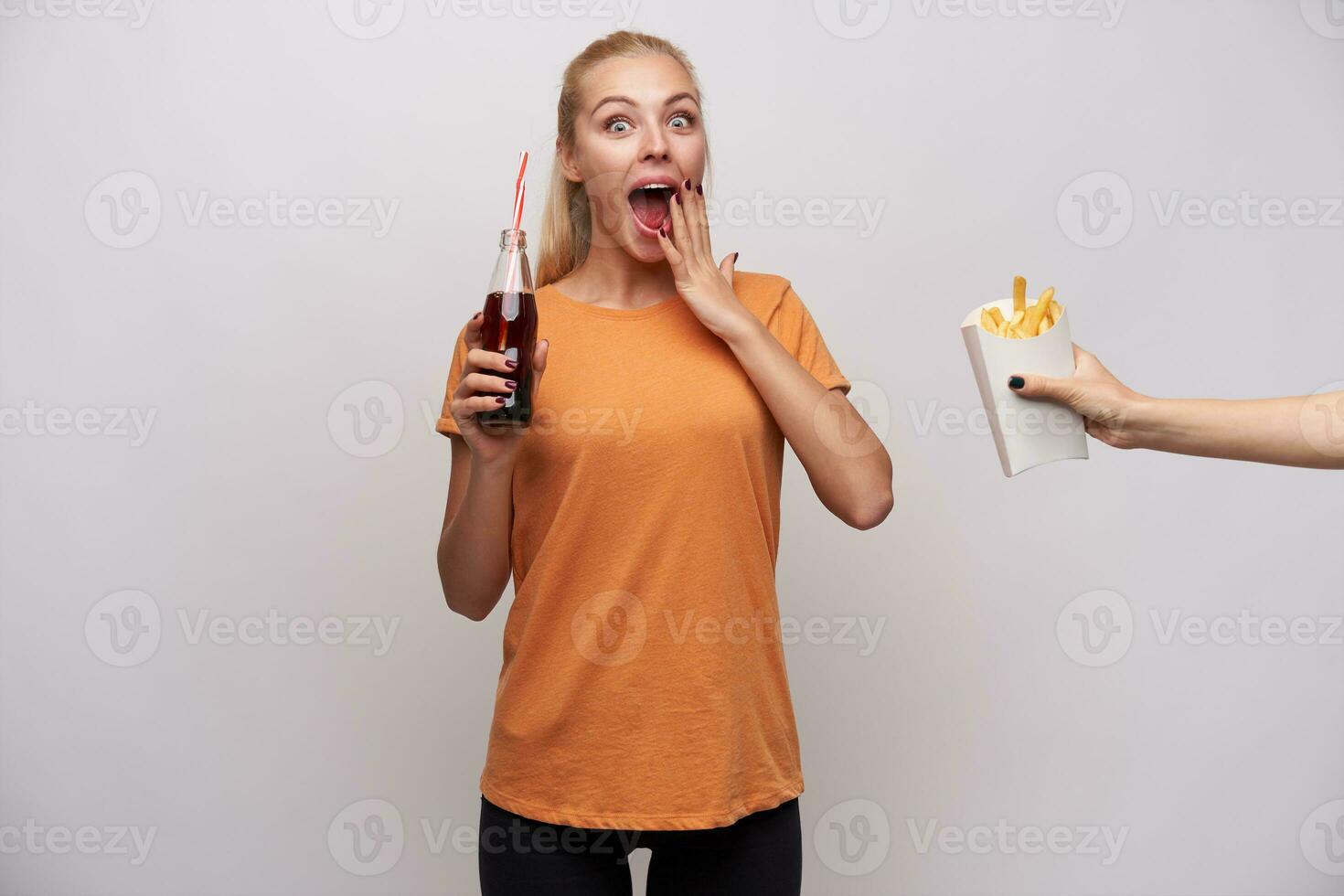 surpreso jovem atraente de olhos azuis Loiras mulher olhando às Câmera com Largo olhos e boca aberto, guardando garrafa do refrigerante dentro mão e ser animado sobre alguém tratando dela francês fritas foto