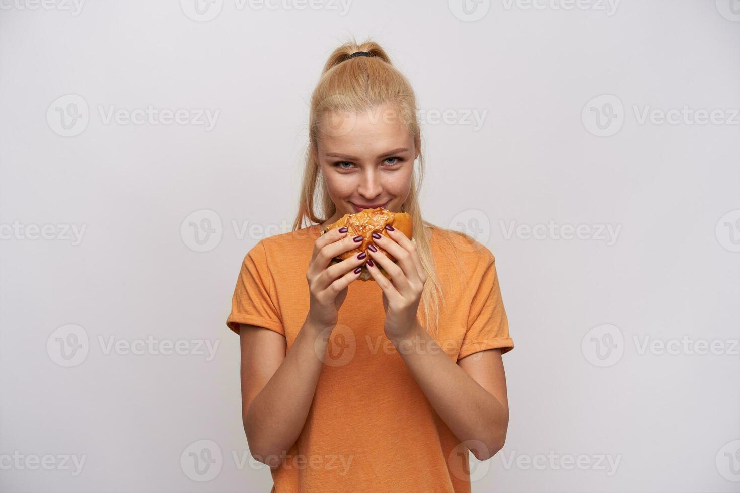 satisfeito jovem atraente Loiras fêmea com rabo de cavalo deliciando fresco hamburguer e olhando alegremente às Câmera, vestindo laranja camiseta enquanto em pé contra branco fundo foto