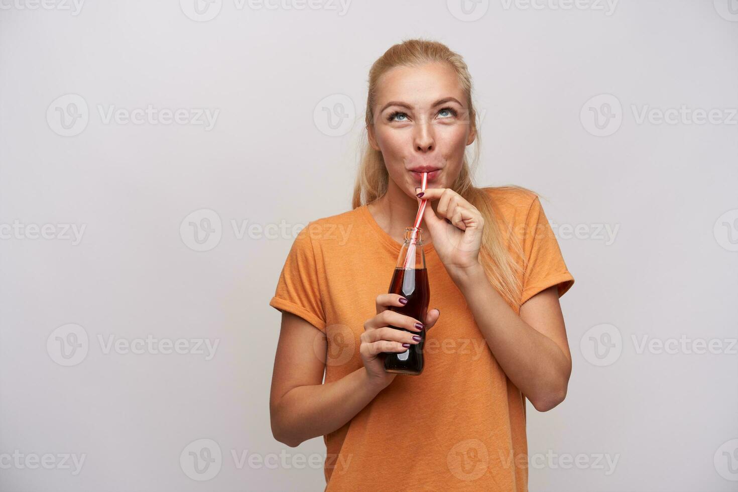 alegre jovem de olhos azuis grandes cabelos Loiras mulher bebendo refrigerante com Palha e olhando positivamente para cima, vestindo casual roupas enquanto em pé sobre branco fundo foto