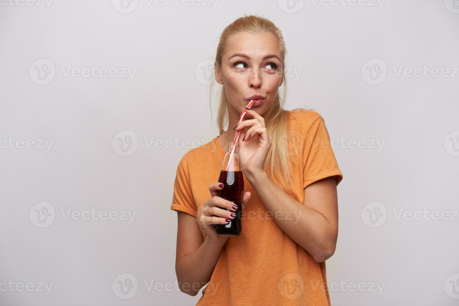 agradável olhando jovem positivo grandes cabelos Loiras mulher com casual Penteado bebendo refrigerante com Palha a partir de vidro garrafa, vestido dentro laranja camiseta enquanto em pé sobre branco fundo foto