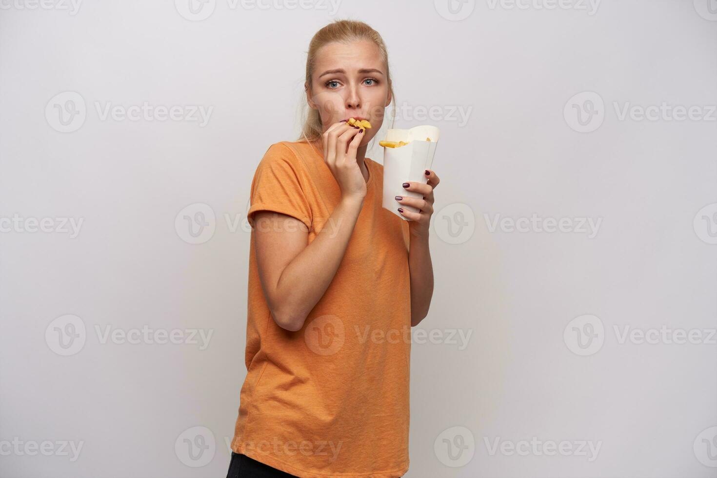 emocional jovem de olhos azuis Loiras mulher com rabo de cavalo Penteado olhando dentro frente do dela e comendo francês fritas enquanto em pé sobre branco fundo dentro laranja camiseta foto