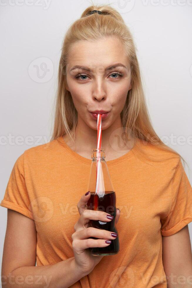 lindo jovem Loiras fêmea com casual Penteado segurando garrafa com refrigerante e bebendo isto com canudo, olhando às Câmera com calma face, vestindo laranja camiseta sobre branco fundo foto