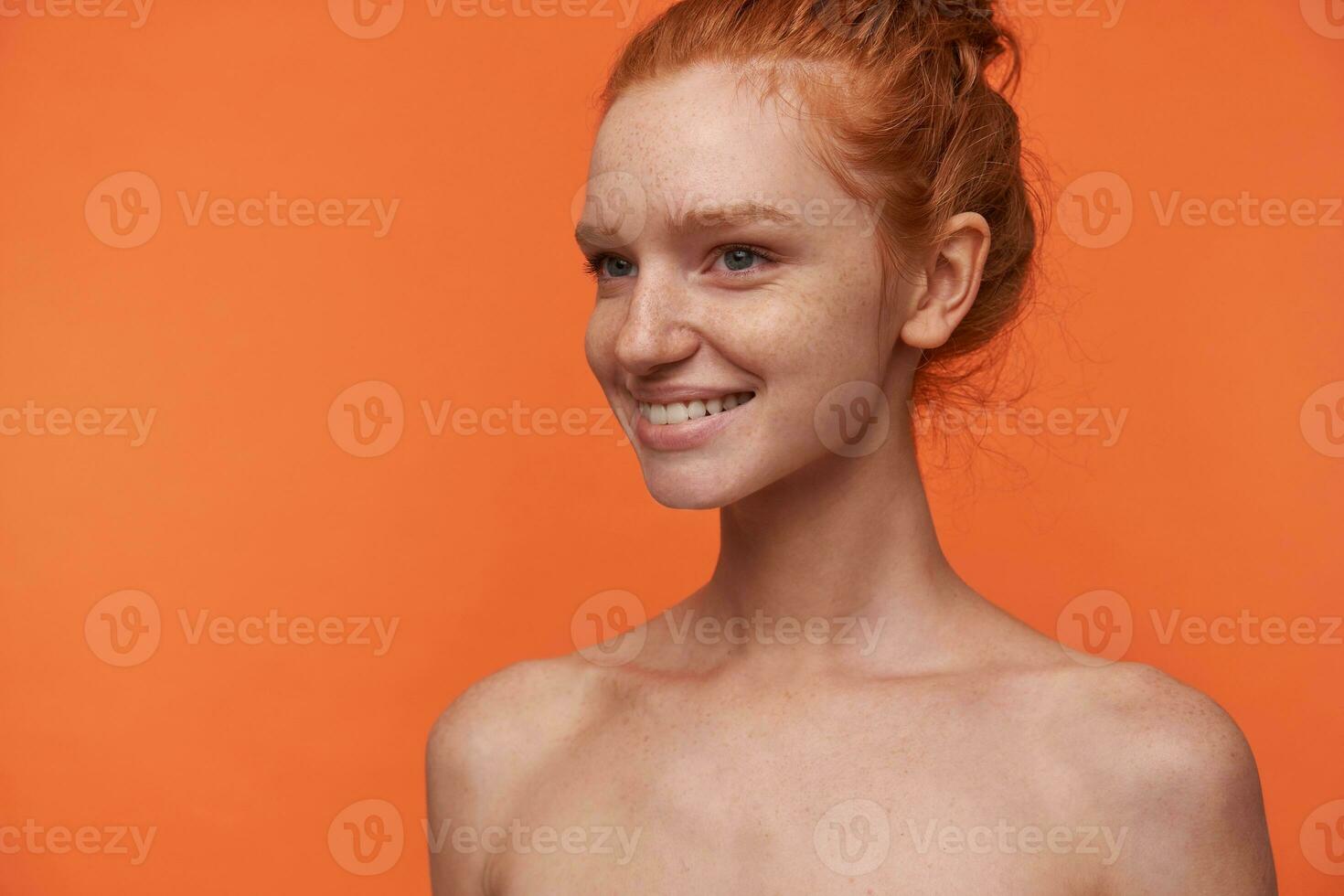 estúdio retrato do jovem atraente cabeça de leitura mulher com pão Penteado isolado sobre laranja fundo, olhando a parte, de lado com encantador sorriso, demonstrando dela branco perfeito dentes foto