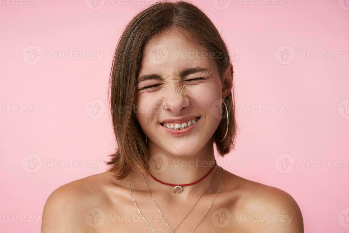 retrato do agradável olhando jovem curto cabelos senhora com natural Maquiagem franzindo a testa dela face enquanto sorridente alegremente com fechadas olhos, isolado sobre Rosa fundo foto