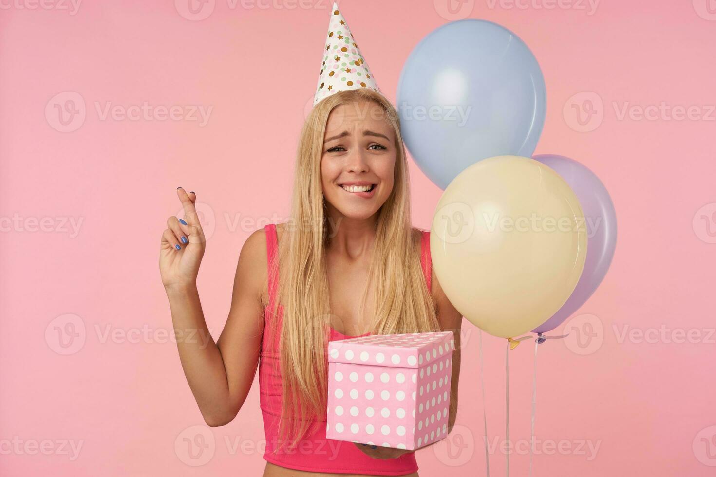 positivo senhora com casual Penteado vestindo Rosa topo e cone chapéu, posando dentro multicolorido ar balões, cruzando dedos para Boa sorte e morder lábio inferior preocupantemente, isolado sobre Rosa fundo foto