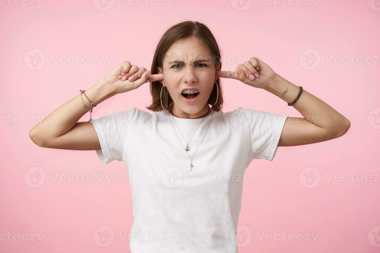 retrato do descontente jovem olhos castanhos curto cabelos fêmea franzindo a testa dela sobrancelhas enquanto olhando às Câmera e fechamento dela orelhas com elevado mãos, isolado sobre Rosa fundo foto