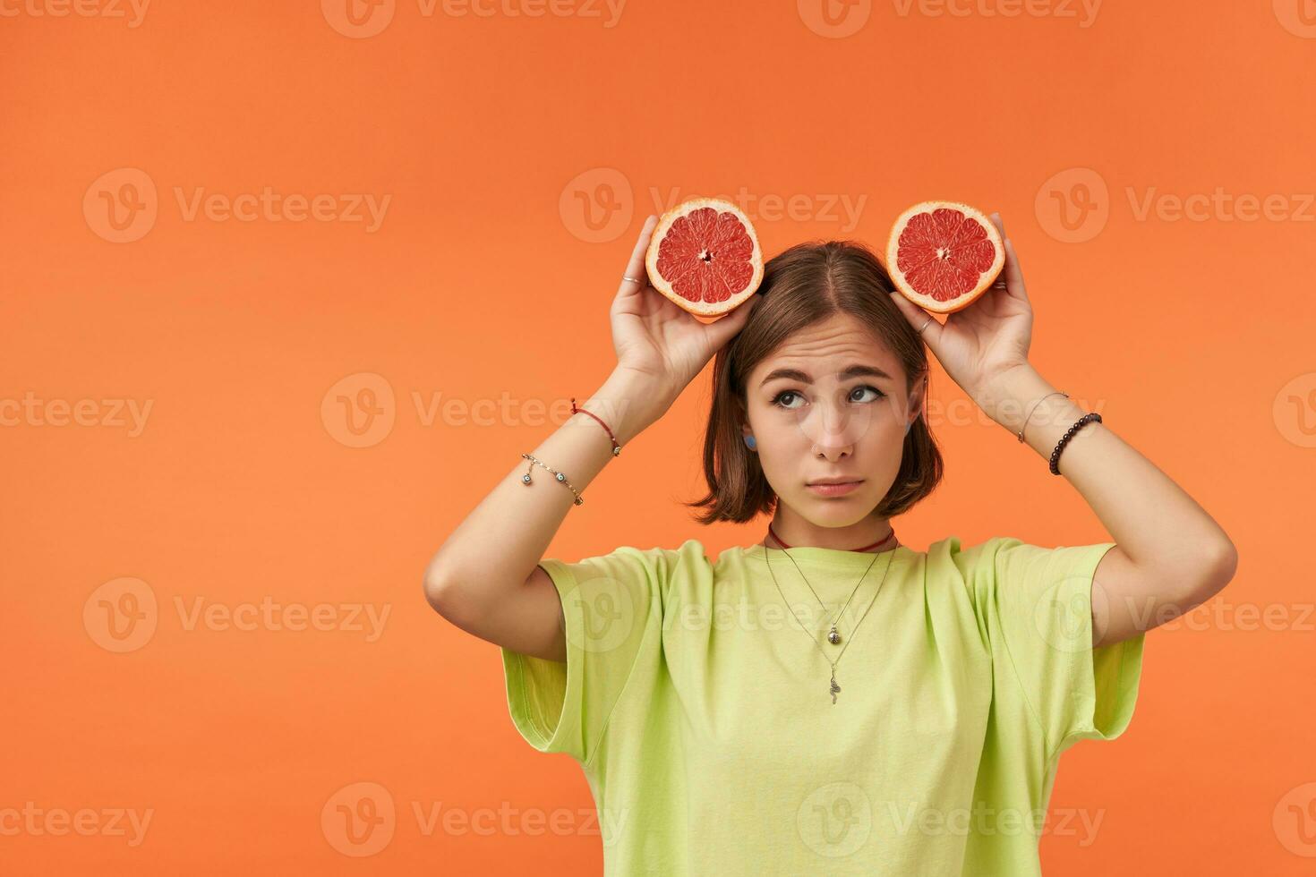 jovem bonita mulher com curto morena cabelo, parece triste, chateado. segurando Toranja sobre dela cabeça. em pé sobre laranja fundo. vestindo verde camiseta, suspensórios e pulseiras foto