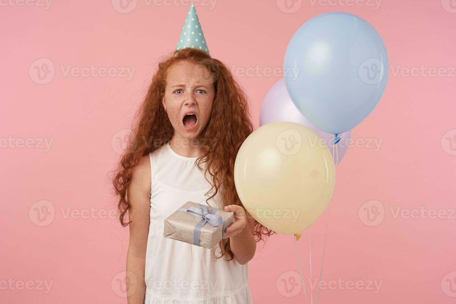 desapontado pequeno menina com grandes foxy cabelo em pé sobre Rosa fundo e gritando tristemente, olhando dentro Câmera e franzindo a testa face com Largo boca aberto, vestindo branco elegante vestir foto