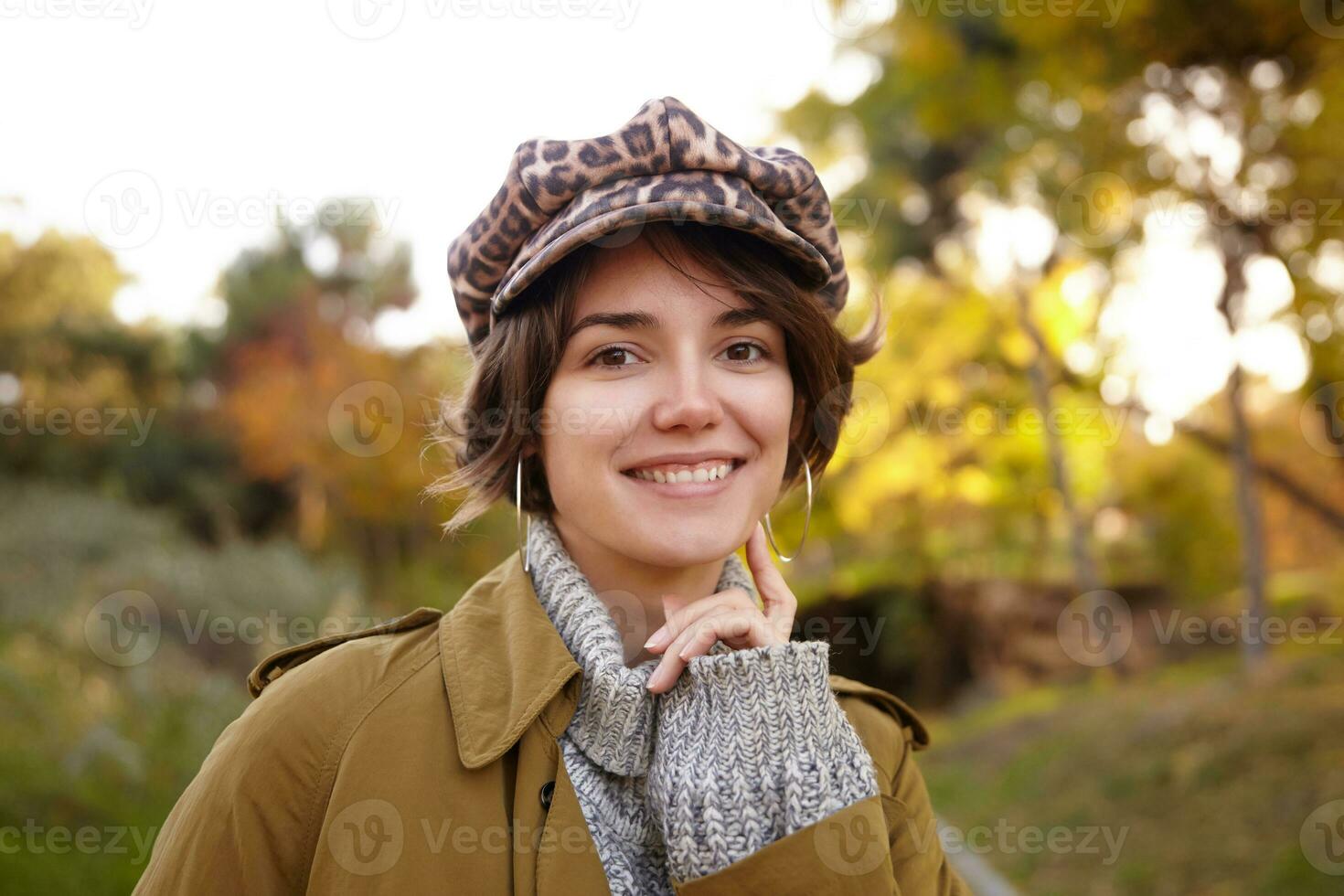 alegre jovem olhos castanhos morena fêmea com casual Penteado olhando positivamente às Câmera com encantador sorrir e tocante dela bochecha com elevado indicador enquanto posando ao ar livre foto