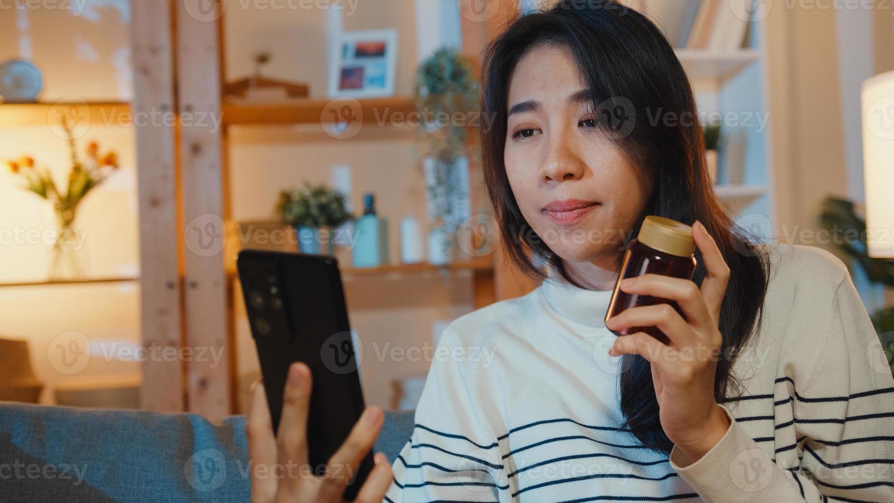 doente jovem asiática segurar medicamento sentar no sofá, videochamada com telefone, consultar o médico em casa à noite. garota toma remédio após ordem do médico, quarentena em casa, vírus de corona de distância social. foto