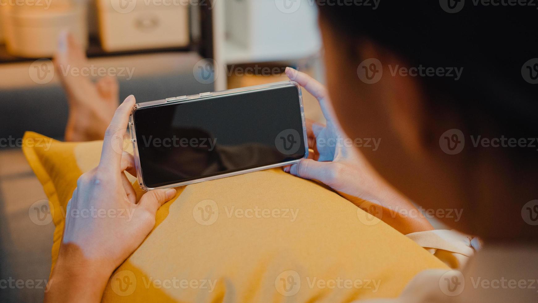 jovem asiática usar telefone inteligente com simulação de tela preta em branco para texto de publicidade, enquanto descansa no sofá na sala de estar em casa à noite moderna. tecnologia chroma key, conceito de design de marketing. foto