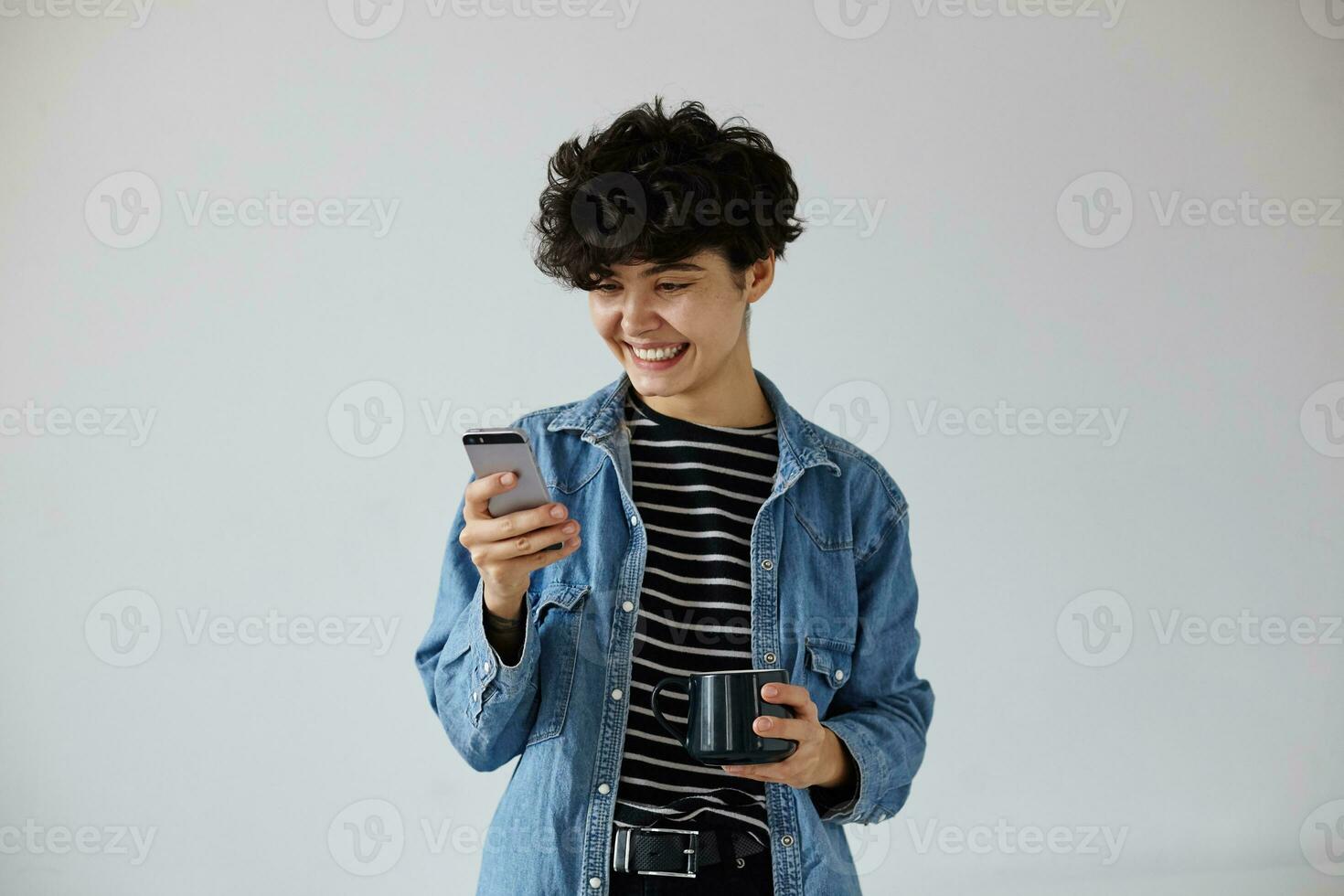 interior foto do encantador jovem positivo Sombrio cabelos encaracolado mulher segurando Smartphone e olhando alegremente em tela, em pé sobre branco fundo com cerâmico copo dentro elevado mão