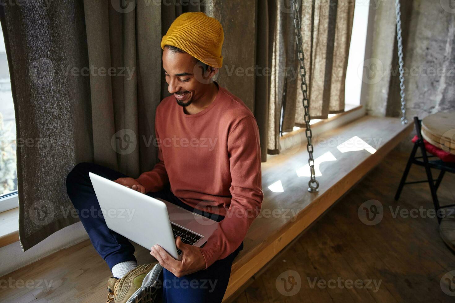 agradável olhando jovem Sombrio esfolado bonita homem dentro mostarda boné sentado sobre grandes de madeira peitoril da janela e guardando computador portátil em dele pernas, olhando às tela alegremente com sincero sorrir foto