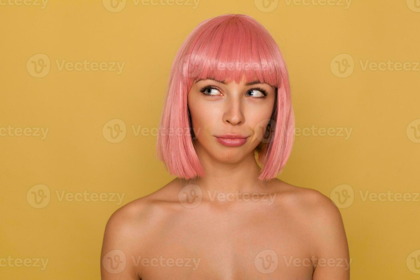 pensativo jovem lindo de olhos azuis fêmea com curto Rosa corte de cabelo guardando dela lábios guardada enquanto olhando pensativamente aparte, plotagem alguma coisa enquanto posando sobre mostarda fundo foto