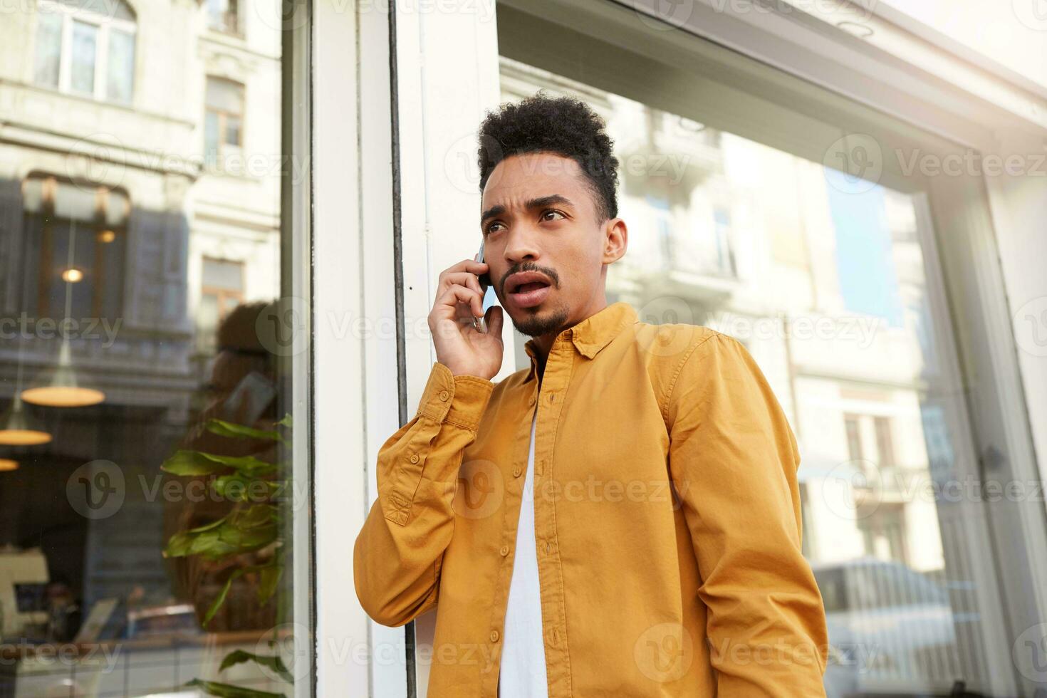 foto do jovem espantado Sombrio esfolado cara dentro amarelo camisa caminhando baixa a rua , fala em a telefone, ouve inacreditável notícias, com Largo aberto boca e olhos, parece atordoado.