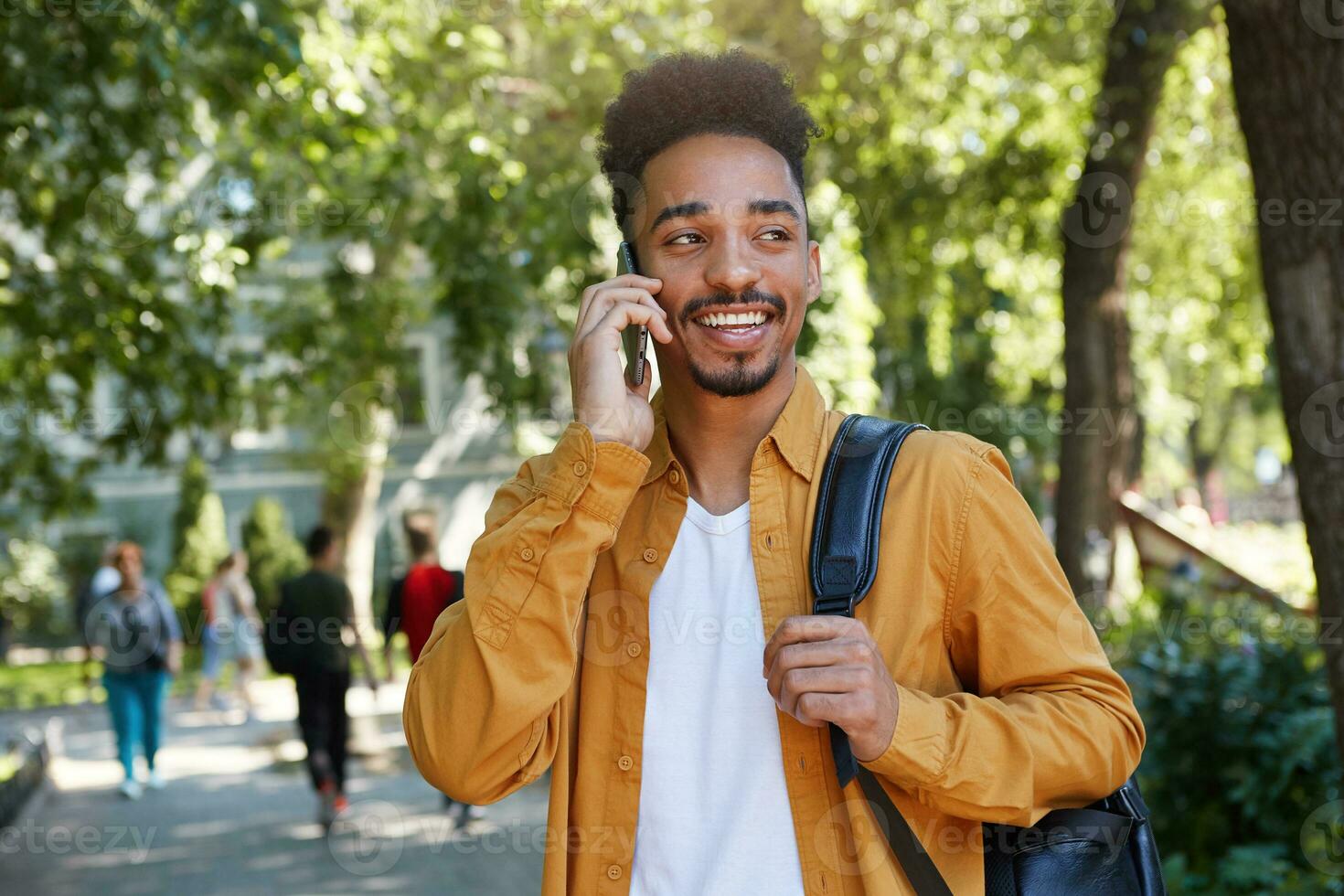 retrato do jovem Sombrio esfolado sorridente cara desgasta dentro uma amarelo camisa e uma branco camiseta com uma mochila em 1 ombro, caminhando dentro a parque e falando em a telefone, sorridente e goza a dia. foto