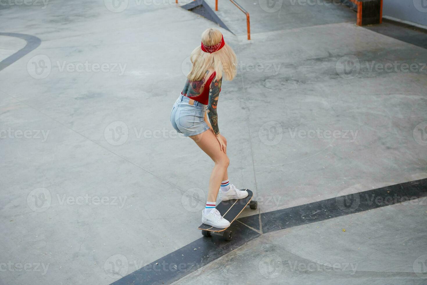 jovem tatuado senhora com Loiras cabelo desgasta dentro uma vermelho camiseta e jeans shorts, com uma tricotado bandana em dela cabeça, invasão em skate, desfrutando a dia dentro patim parque. foto