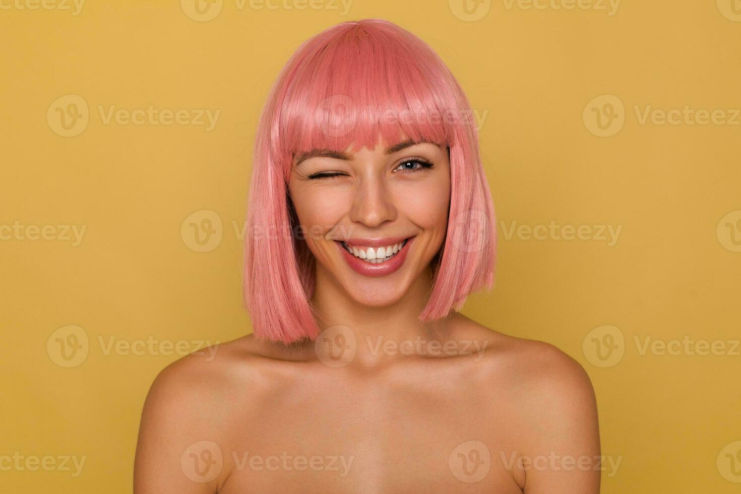 fechar-se do encantador alegre jovem Rosa cabelos senhora com curto na moda corte de cabelo posando sobre mostarda fundo, olhando de brincadeira às Câmera e dando piscadela foto