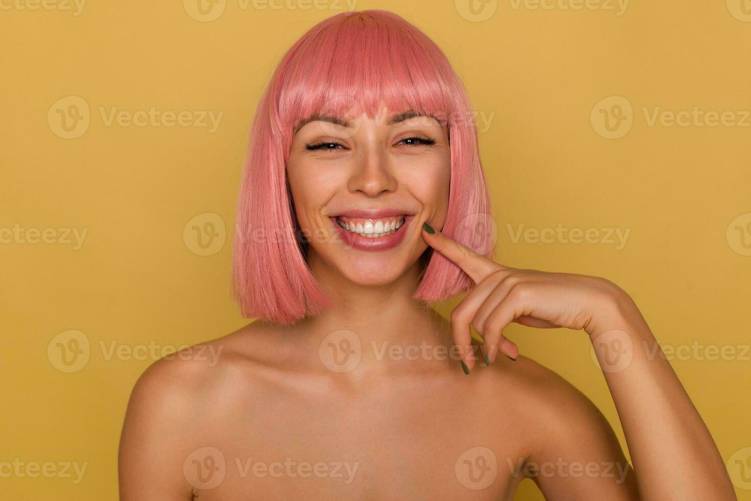 alegre jovem atraente senhora com curto Rosa corte de cabelo mostrando dela branco perfeito dentes enquanto sorridente amplamente às Câmera, tocante dela face com elevado índice dedo enquanto posando sobre mostarda fundo foto