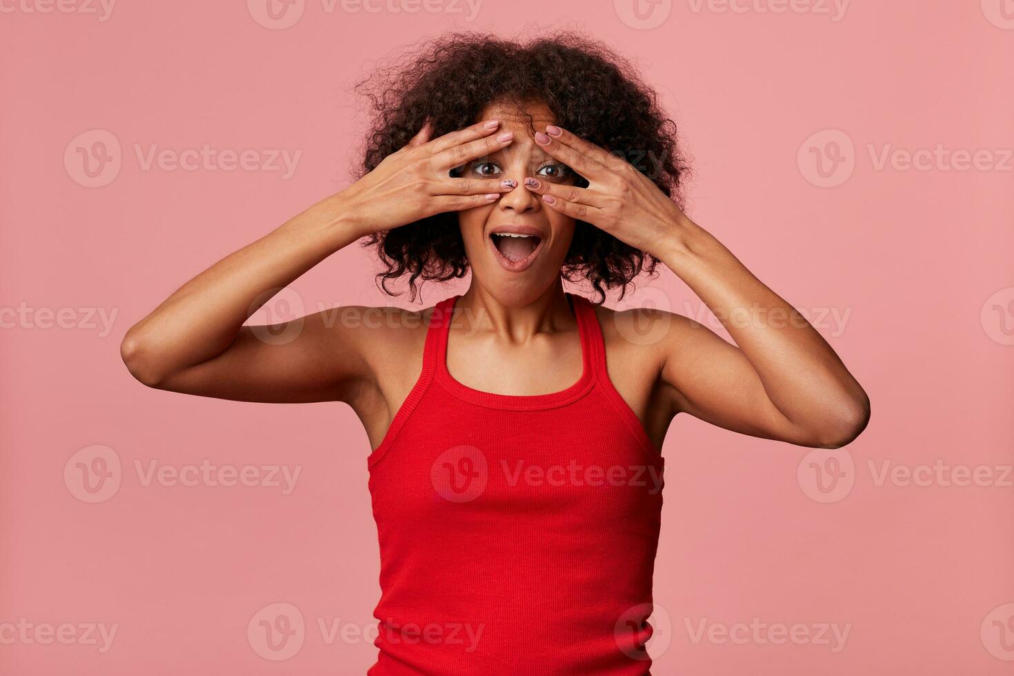 estúdio foto do engraçado jovem beleza africano americano senhora com encaracolado Sombrio cabelo, vestindo vermelho camiseta, olhando através dedos com Largo aberto boca e olhos. isolado sobre oink fundo.