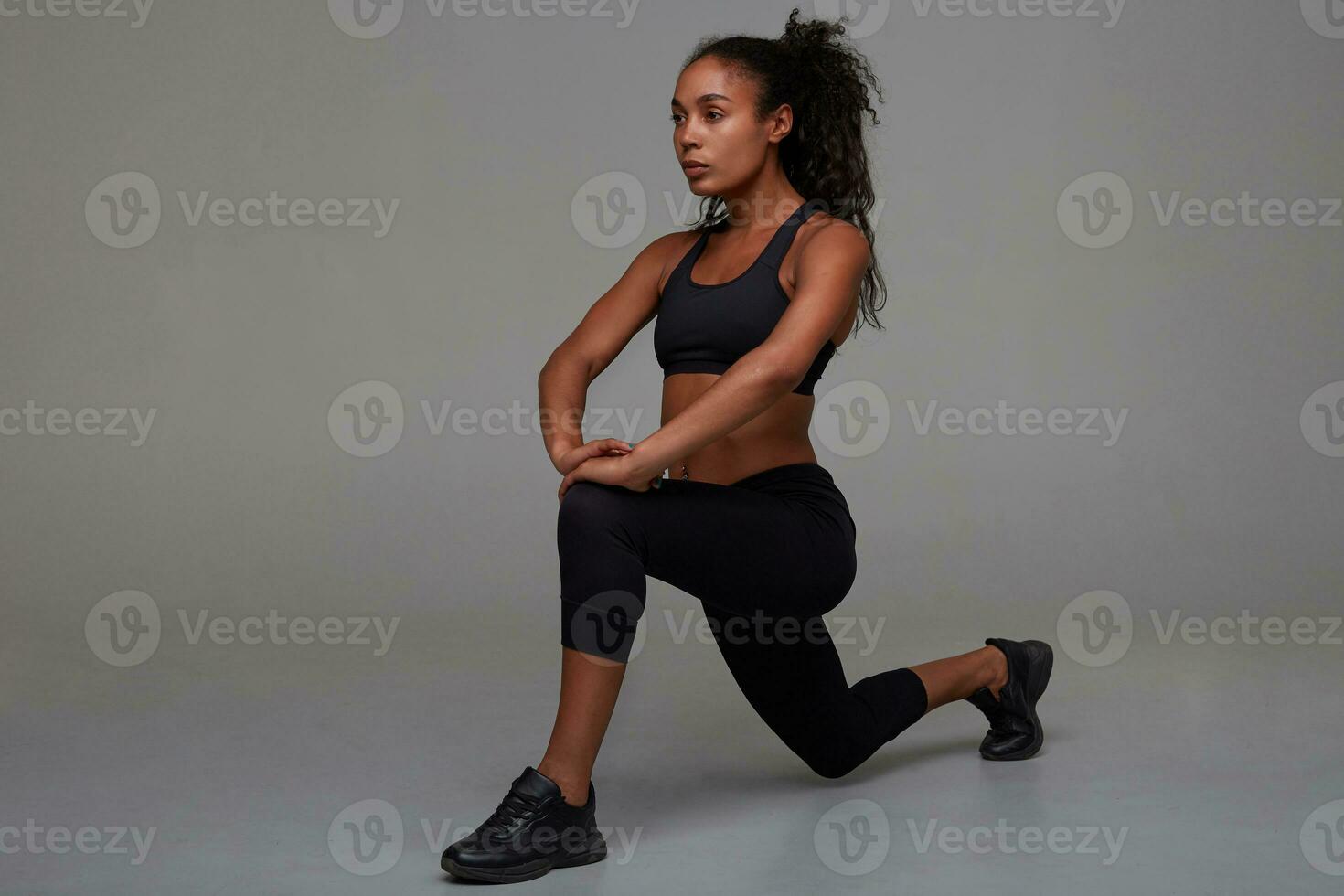 tiro do jovem lindo Sombrio esfolado morena senhora com rabo de cavalo Penteado alongamento músculos depois de ginástica sessão, vestido dentro Preto desportivo topo e leggins sobre cinzento fundo foto