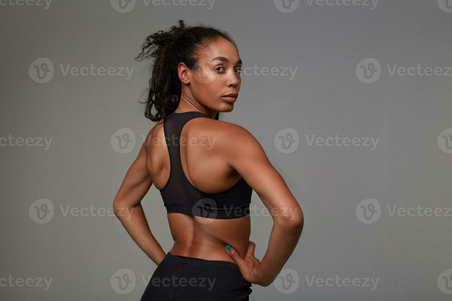 estúdio tiro do jovem encaracolado morena fêmea com Sombrio pele olhando sobre dela ombro enquanto posando sobre cinzento fundo dentro Atlético Preto roupas. ginástica masculino modelo foto