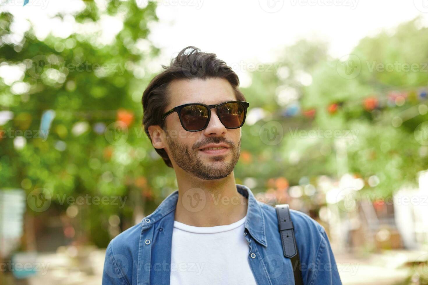 ao ar livre tiro do lindo barbudo jovem homem dentro oculos de sol posando sobre cidade jardim em caloroso ensolarado dia, vestindo azul camisa e branco camiseta foto