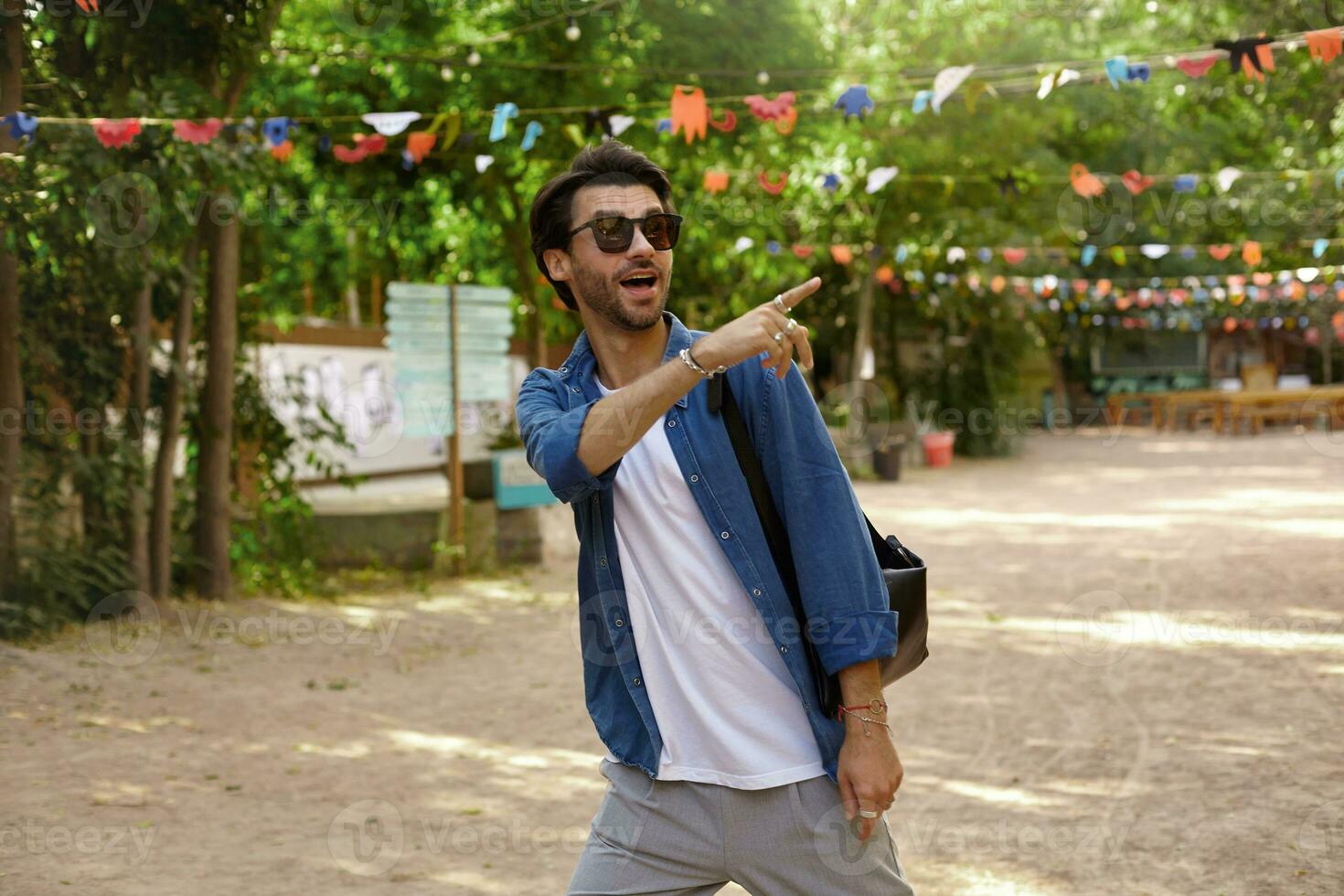 surpreso bonita jovem homem com barba caminhando ao longo verde parque dentro casual vestem e oculos de sol, percebendo alguma coisa interessante e apontando lá com indicador foto