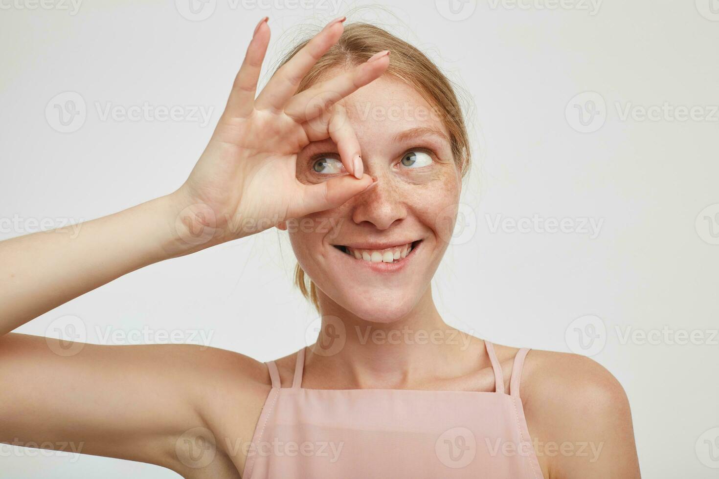 interior tiro do alegre jovem adorável ruiva fêmea morder positivamente dela lábio inferior e guardando elevado Está bem placa em dela face enquanto em pé sobre branco fundo foto