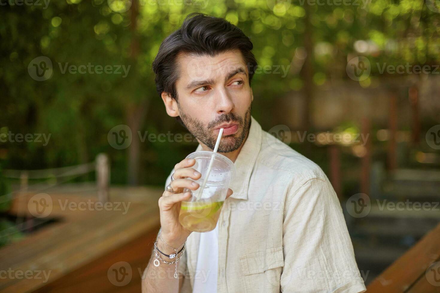 ao ar livre retrato do encantador jovem barbudo masculino com Sombrio cabelo olhando a parte, de lado e bebendo limão com canudo, posando sobre verde cidade jardim foto