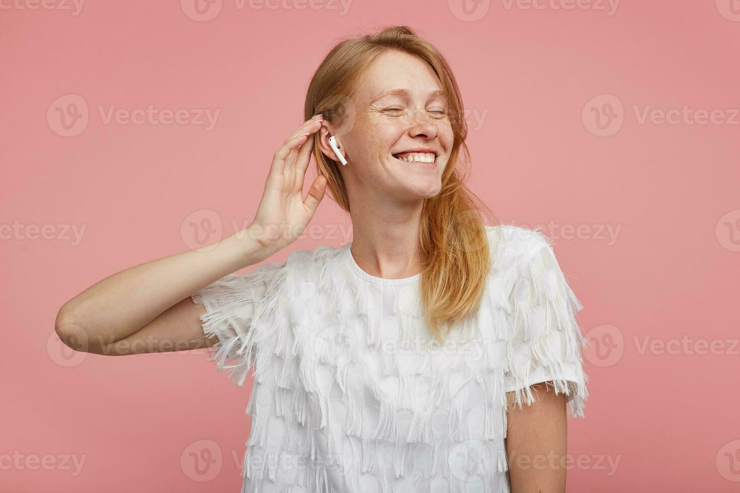 Boa olhando jovem feliz ruiva mulher com natural Maquiagem levantando mão para dela cabeça enquanto posando sobre Rosa fundo, guardando olhos fechadas enquanto desfrutando música rastrear dentro dela fones de ouvido foto