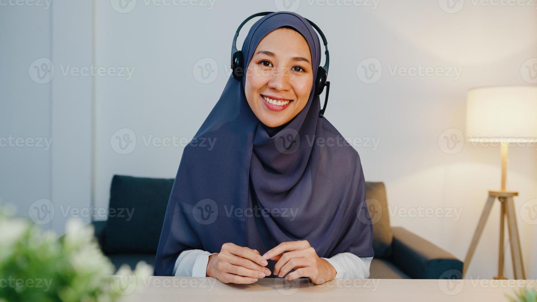 A senhora muçulmana asiática usa fone de ouvido usando laptop e fala com colegas sobre o relatório de venda em uma videoconferência enquanto trabalha em casa à noite. distanciamento social, quarentena para o vírus corona. foto