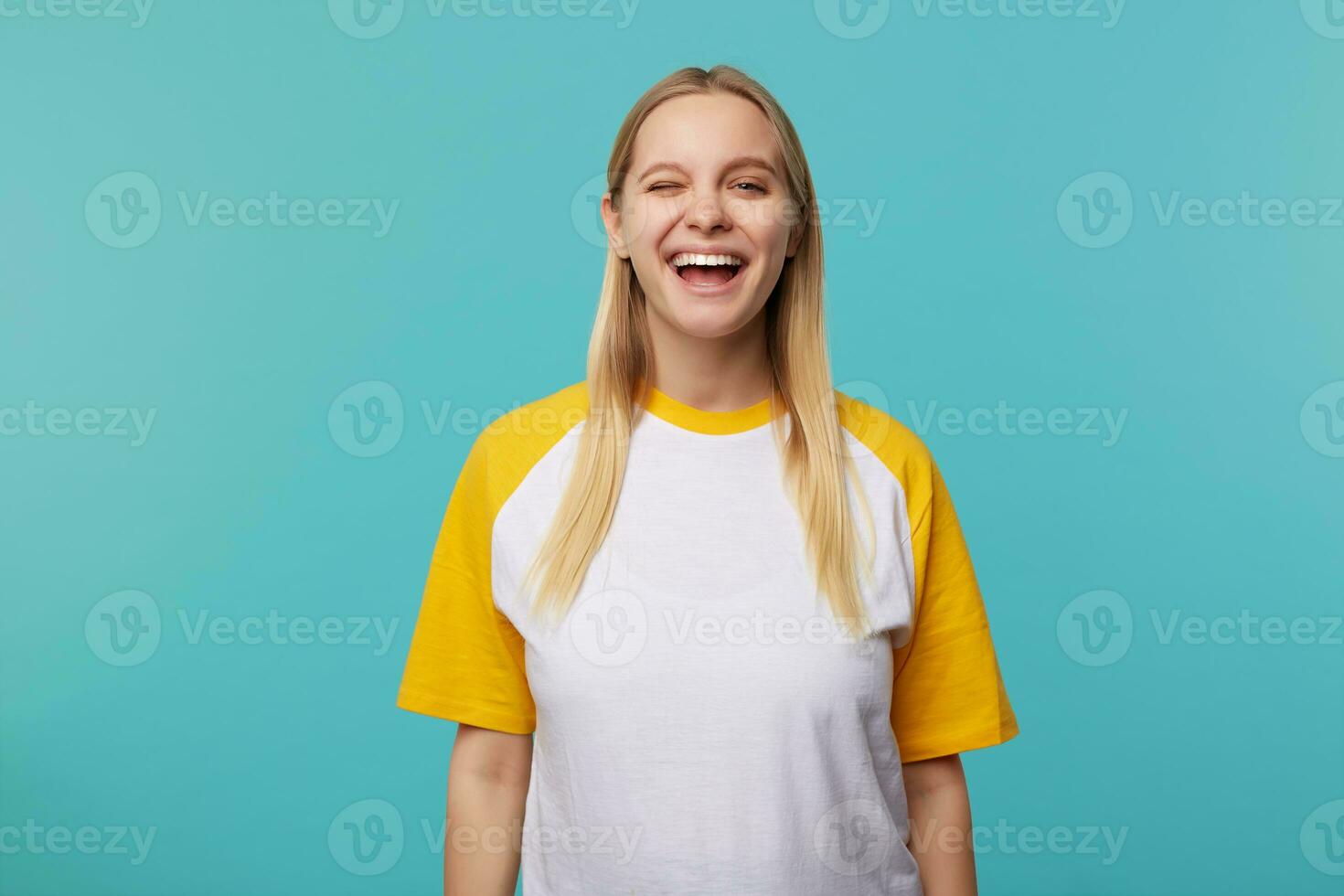 interior foto do alegre jovem bonita grandes cabelos Loiras mulher piscando alegremente às Câmera e sorridente amplamente enquanto em pé sobre azul fundo com mãos baixa