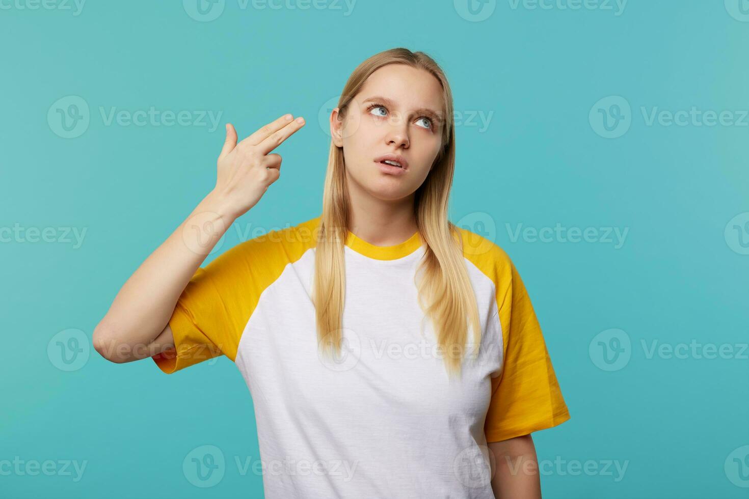 descontentamento jovem bonita louro mulher com natural Maquiagem levantando mão com arma gesto para dela cabeça e olhando para cima com entediado face, isolado sobre azul fundo foto