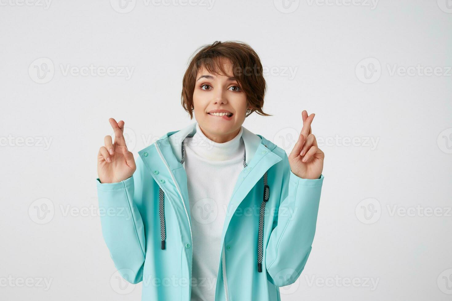 retrato do positivo jovem agradável senhora dentro azul chuva casaco, parece às a Câmera com feliz expressões, com cruzado dedos e fechadas olhos, esperanças para sorte e mordidas lábio, carrinhos sobre branco muro. foto