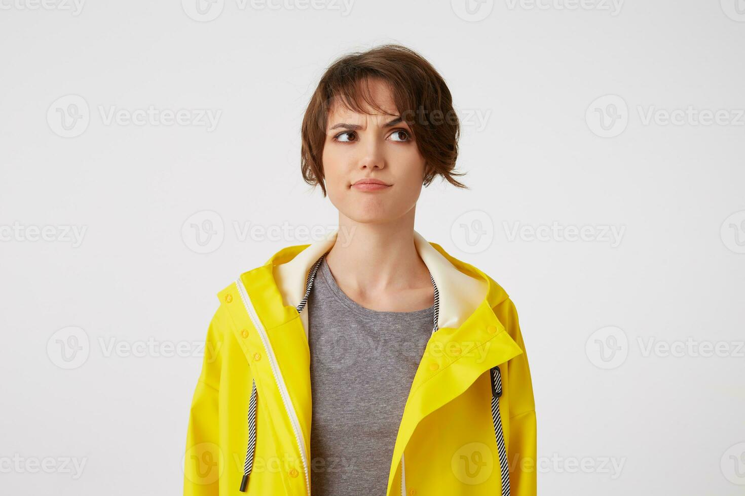 jovem duvidando cabelo curto mulher dentro chuva amarela casaco, franzindo a testa parece ausente, carrinhos sobre branco fundo, parece descontentamento e duvidando. foto