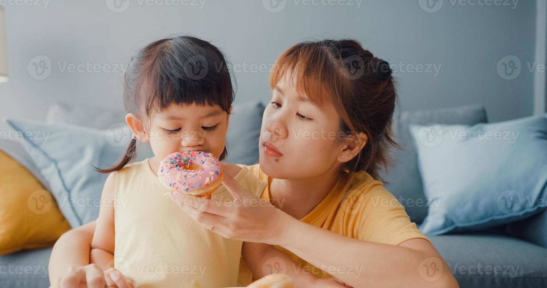 feliz alegre família asiática mãe e criança menina comendo donuts e se divertindo, relaxar desfrutar no sofá na sala de estar em casa. passar um tempo juntos, distância social, quarentena para coronavírus. foto